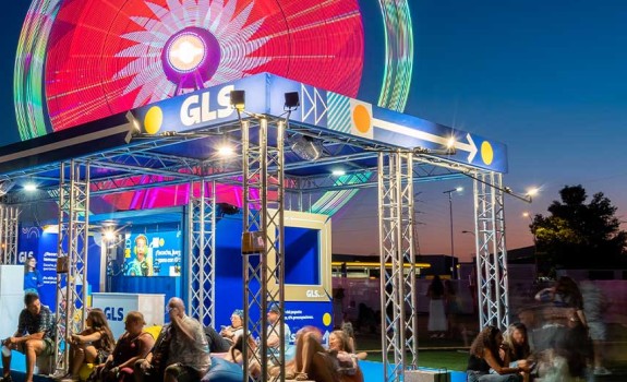 Stand nocturno de GLS iluminado en Mad Cool Festival, con asistentes descansando y noria iluminada de fondo