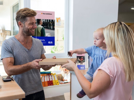 A woman holding her daughter hands a package to a man working in a parcel shop